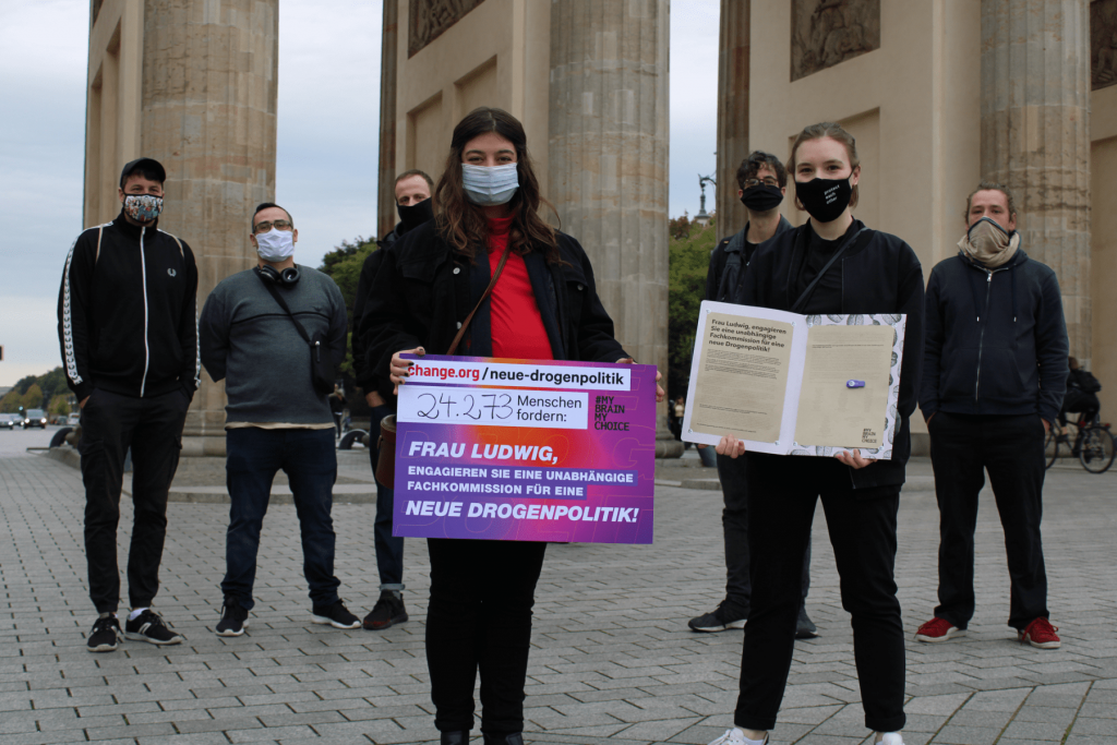 Zhana Jung und Philine Edbauer präsentieren vor dem Brandenburger Tor ein Schild mit der Forderung der Petition und der Zahl 24.273 Unterschriften sowie den ausgedruckten Petitionstext mit den Erstunterzeichner*innen